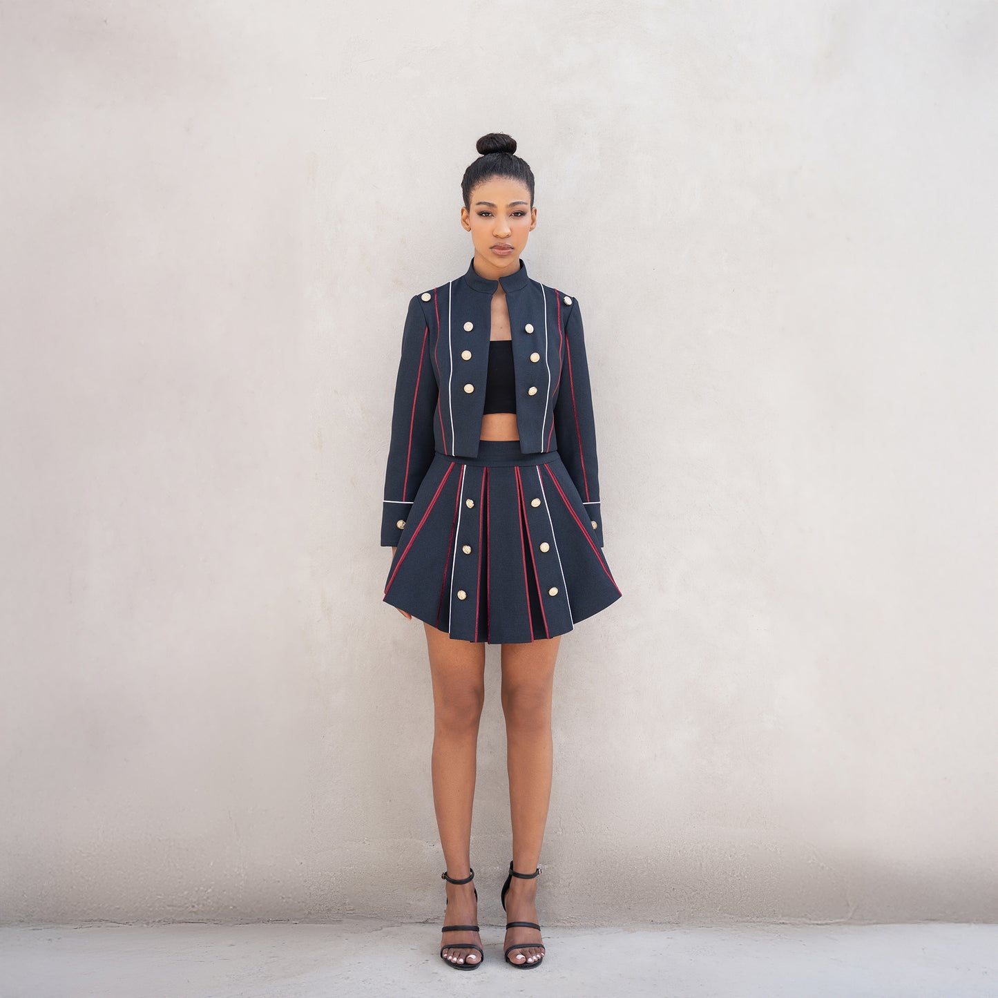 A woman wearing the Axis set, consisting of a cropped Fastoni jacket with golden buttons and a pleated skirt with red and white laser-cut lines.